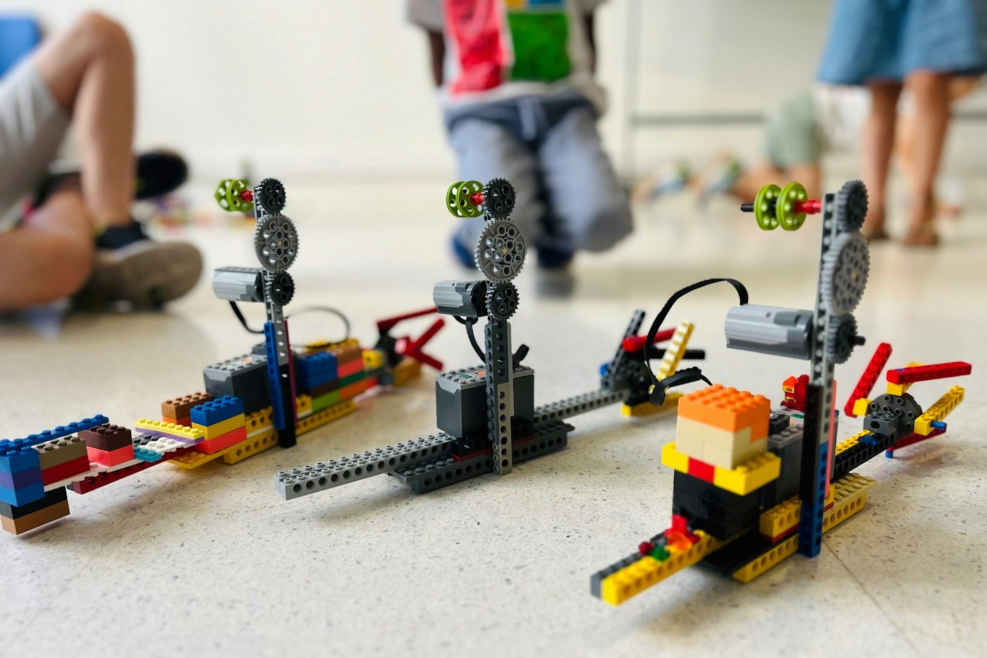 Three robotic models built from plastic bricks on a light floor with children nearby